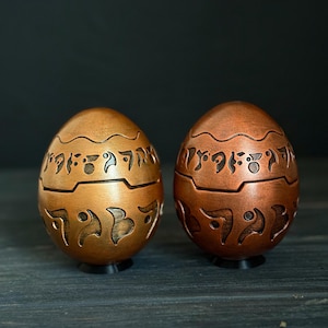Peut inclure: Deux œufs décoratifs avec des sculptures complexes et une finition cuivrée. Les œufs sont sur une surface en bois foncé.