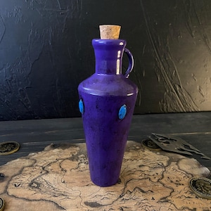 Könnte beinhalten: Eine tiefviolette Keramikflasche mit Korken und kleinem Griff. Die Flasche ist mit zwei ovalen Akzenten in Türkis verziert. Die Flasche wird auf einer Karte mit Goldmünzen präsentiert.