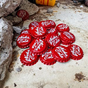 May include: A pile of red bottle caps, each bearing the white text "Nuka Cola". The caps are arranged on a distressed surface, with a yellow battery and other items visible. The caps are a bright red colour.