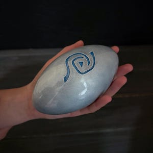 Puede incluir: Una piedra lisa, ovalada, de color azul claro con un diseño de espiral azul. La piedra se sostiene en una mano sobre un fondo oscuro. La superficie de la piedra tiene un brillo sutil y nacarado.