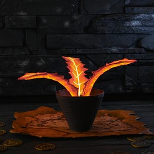 May include: A decorative, glowing plant with orange and yellow leaves in a black pot. The pot sits on a map-shaped tray, surrounded by gold coins, against a dark background. The plant's leaves have a bright, fiery appearance.
