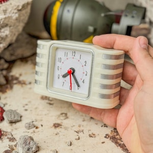 May include: A cream-colored retro alarm clock with silver accents and a red second hand. The clock has a square face with black numerals and is held against a backdrop of a bomb and rubble.