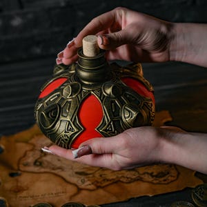 May include: A decorative, round bottle with a red interior and ornate, bronze-colored metalwork. The bottle has a cork stopper and is held over a map and several bronze coins. The bottle's design suggests a fantasy or medieval theme.