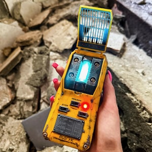 May include: A yellow handheld electronic device with a weathered finish, featuring a glowing blue light, a flip-up blue grid top, and a metal plate with technical text including the word "ВІДГУК". It has black switches and a small red indicator light.
