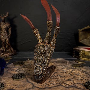 May include: A detailed, bronze-colored claw prop with three long, red-tipped claws. The base features three circular medallions. The prop is set on a map with scattered coins, suggesting a fantasy or role-playing theme.