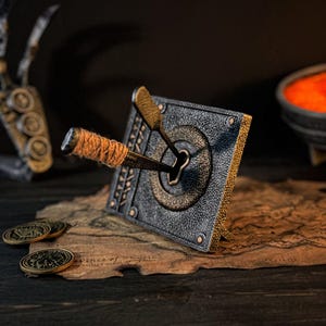 May include: A detailed, antique-style lock with a key inserted, set against a dark background. The lock is square, with a bronze-colored key and accents. The key handle is wrapped in orange twine. Two bronze coins and a map are in the foreground.