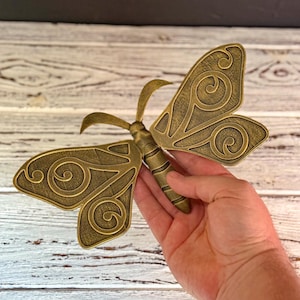 Puede incluir: Una mariposa de metal de color dorado con intrincados patrones en espiral en sus alas. La mariposa está sostenida en una mano, con el cuerpo de la mariposa apuntando hacia el espectador.