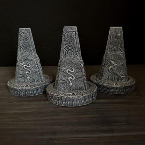 May include: Three grey, stone-like decorative objects with a weathered appearance. Each has a conical shape with carved spiral and animal designs, including a snake and a fish. The objects sit on a circular base, creating a unique, ancient aesthetic.