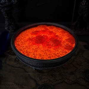 May include: A dark, round, textured bowl filled with glowing orange crystals, resembling lava. The bowl sits on a map with a coin and statues in the background. The crystals emit a bright, warm light.