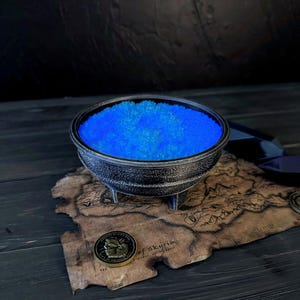 May include: A dark, metallic cauldron filled with bright blue crystals, set on a map with a coin. The cauldron has three legs and a detailed rim. The map and coin add a thematic element to the scene.