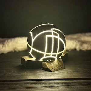 May include: A black sphere with white lines, resembling a globe, sits on a bronze base with the inscription "Apples of Eden". The sphere is illuminated from within, casting a warm glow.