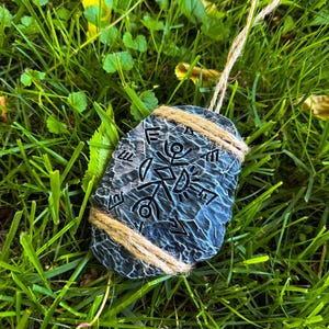 May include: A dark blue, stone-like pendant with etched symbols, wrapped with twine and a twine loop for hanging. The pendant is set against a backdrop of green grass.