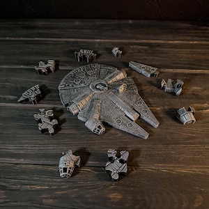 May include: A detailed, grey model of the Millennium Falcon spaceship, with its separate components, arranged on a dark wooden surface. The model features intricate detailing and a weathered appearance, suggesting a vintage or collectible item.