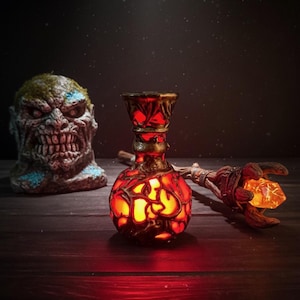 Könnte beinhalten: Eine leuchtende, verzierte rote und goldene Glasflasche mit passendem Stab und einer Monsterkopf-Statue. Die Flasche hat ein Gittermuster, und der Stab hält einen leuchtenden orangefarbenen Kristall. Der Monsterkopf ist grün und blau.