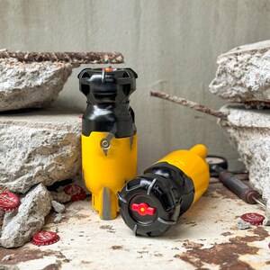 May include: Two bottle stoppers with a black and yellow design, resembling a miniature bomb. One is upright, the other is lying on its side. The stoppers have a red accent and are set against a textured background.