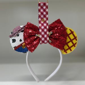 Può includere: Una fascia decorativa con un fiocco rosso con paillettes, una fascia bianca e due decorazioni circolari per le orecchie. Un orecchio è bianco con macchie nere, l'altro è giallo con un motivo a griglia. È attaccato un cinturino a scacchi rosso e bianco con una chiusura dorata.