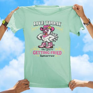 Puede incluir: Una camiseta de color verde claro con el gr&aacute;fico de un pollo blanco que lleva un sombrero de vaquero rosa, un pa&ntilde;uelo rosa y botas de vaquero rosas. El texto dice LIVE TODAY en negro y GETTING FRIED tomorrow en rosa, con adornos amarillos.