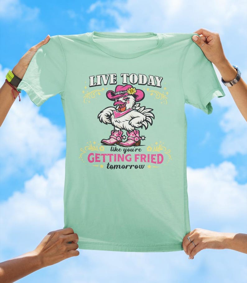 Puede incluir: Una camiseta de color verde claro con el gr&aacute;fico de un pollo blanco que lleva un sombrero de vaquero rosa, un pa&ntilde;uelo rosa y botas de vaquero rosas. El texto dice LIVE TODAY en negro y GETTING FRIED tomorrow en rosa, con adornos amarillos.
