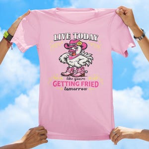 Puede incluir: Una camiseta rosa claro con un gr&aacute;fico de un pollo blanco que lleva un sombrero de vaquero rosa, un pa&ntilde;uelo rosa y botas de vaquero rosas. El texto dice LIVE TODAY like you're GETTING FRIED tomorrow en letras negras y rosas.