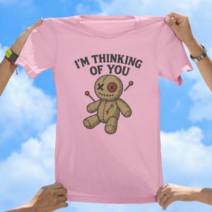 May include: Pink t-shirt featuring a voodoo doll graphic and the text "I'm thinking of you".