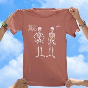 May include: A terracotta-colored t-shirt with a graphic of two skeletons. One skeleton has a slice of pizza in its ribcage and the other does not. The text on the shirt reads "Did you eat my pizza?" and "No."
