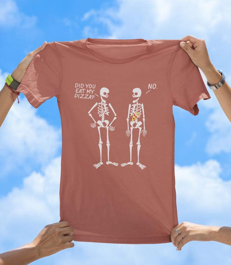 May include: A terracotta-colored t-shirt with a graphic of two skeletons. One skeleton has a slice of pizza in its ribcage and the other does not. The text on the shirt reads "Did you eat my pizza?" and "No."