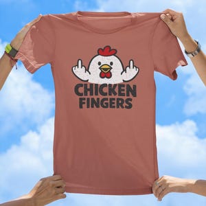 May include: A terracotta-coloured t-shirt featuring a graphic of a white chicken with a red comb and yellow beak, displaying both middle fingers. Below the chicken, the text CHICKEN FINGERS is printed in a bold, distressed black font.