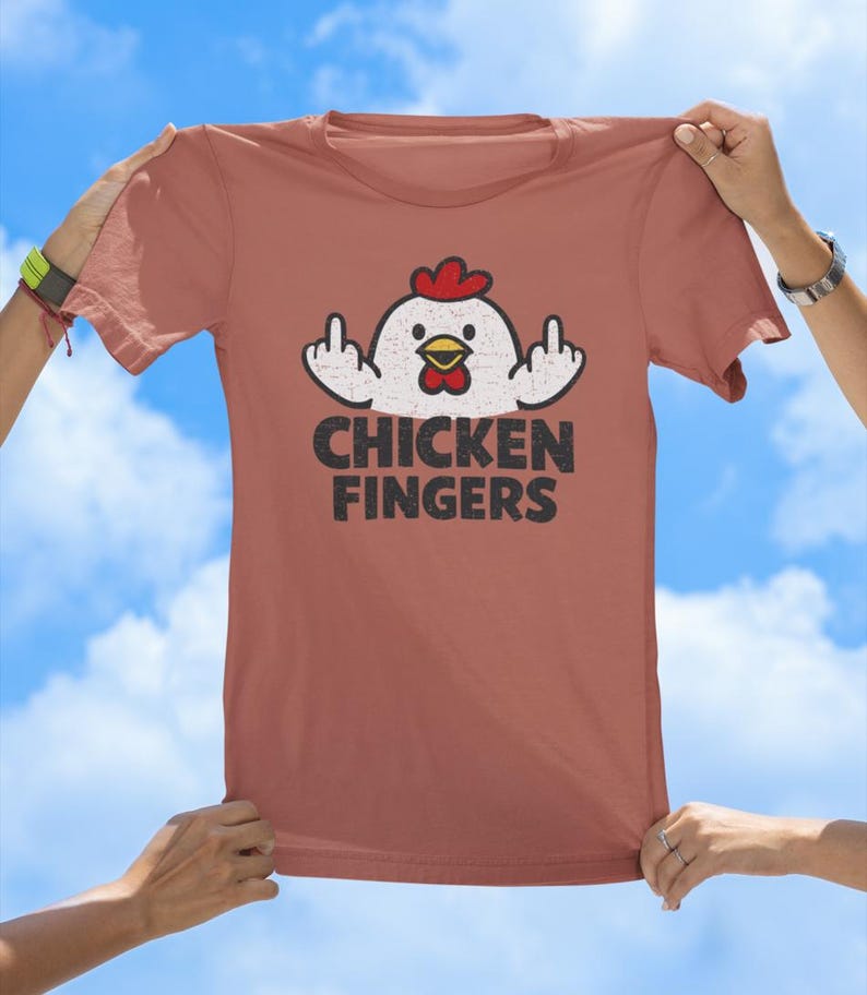 May include: A terracotta-coloured t-shirt featuring a graphic of a white chicken with a red comb and yellow beak, displaying both middle fingers. Below the chicken, the text CHICKEN FINGERS is printed in a bold, distressed black font.
