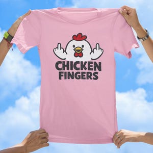May include: A light pink t-shirt featuring a graphic of a white chicken with a red comb and yellow beak, displaying middle fingers on both hands. Below the chicken, the text CHICKEN FINGERS is printed in a distressed black font.