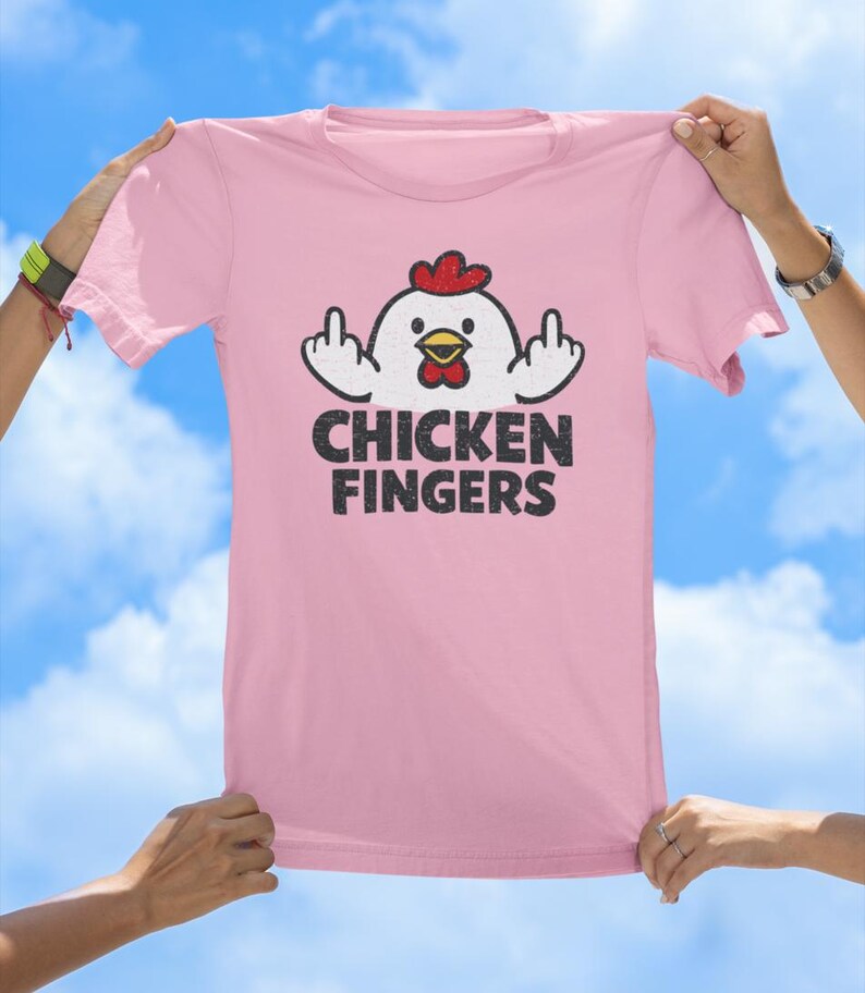 May include: A light pink t-shirt featuring a graphic of a white chicken with a red comb and yellow beak, displaying middle fingers on both hands. Below the chicken, the text CHICKEN FINGERS is printed in a distressed black font.
