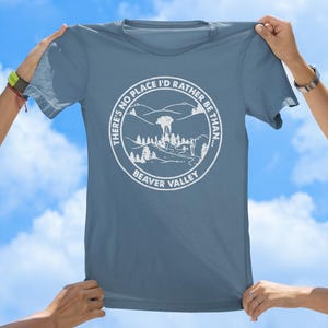 May include: A blue t-shirt with a white circular graphic featuring a mountain landscape and the text "THERE'S NO PLACE I'D RATHER BE THAN... BEAVER VALLEY". The shirt is held up against a blue sky.