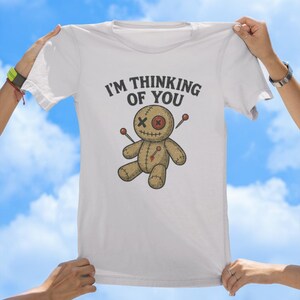 May include: White t-shirt featuring the text "I'm thinking of you" and a cartoon voodoo doll with needles sticking out of it.