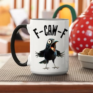 May include: White coffee mug with a black rim and handle. The mug features a black cartoon crow with its mouth open and the text "F-CAW-F" in black.