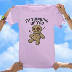 May include: A pink t-shirt featuring the text "I'm thinking of you" and a cartoon voodoo doll with needles sticking out of it.