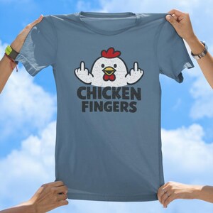 May include: A slate blue short-sleeved t-shirt featuring a graphic of a white chicken with a red comb and wattle. The chicken is shown with its wings raised, displaying middle fingers. Below the graphic, the text reads CHICKEN FINGERS in black.