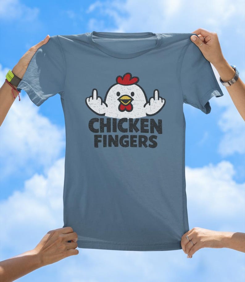 May include: A slate blue short-sleeved t-shirt featuring a graphic of a white chicken with a red comb and wattle. The chicken is shown with its wings raised, displaying middle fingers. Below the graphic, the text reads CHICKEN FINGERS in black.