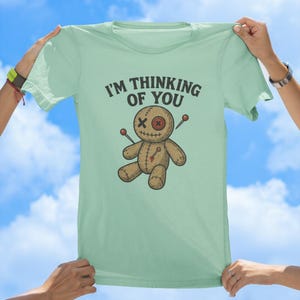 May include: A light green t-shirt featuring a cartoon voodoo doll and the text "I'm thinking of you".