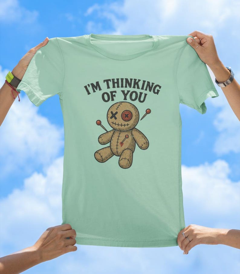 May include: A light green t-shirt featuring a cartoon voodoo doll and the text "I'm thinking of you".