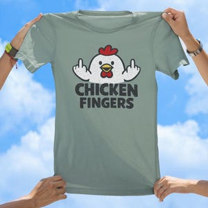 May include: A sage green t-shirt featuring a graphic of a white chicken with a red comb and yellow beak, holding up both middle fingers. Below the chicken, the text CHICKEN FINGERS is printed in a bold, distressed black sans-serif font.