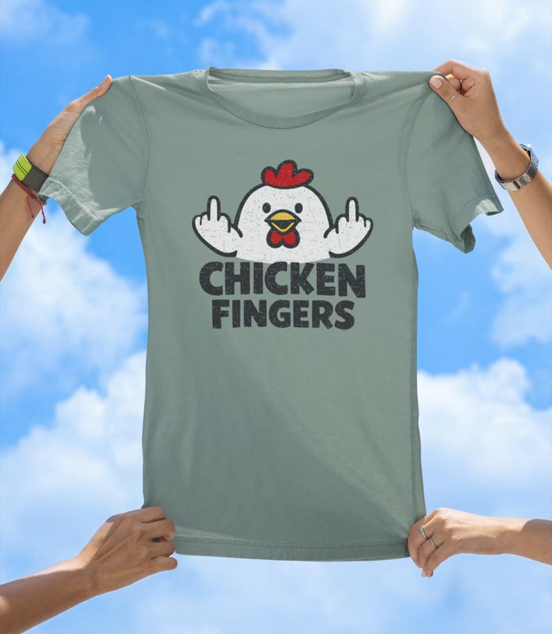May include: A sage green t-shirt featuring a graphic of a white chicken with a red comb and yellow beak, holding up both middle fingers. Below the chicken, the text CHICKEN FINGERS is printed in a bold, distressed black sans-serif font.