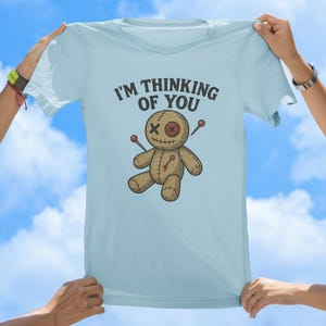 May include: Light blue t-shirt featuring a voodoo doll graphic and the text "I'm thinking of you".