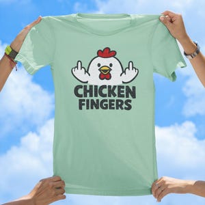 May include: A light green t-shirt featuring a graphic of a white chicken with a red comb and yellow beak, holding up its middle fingers on both sides. Below the chicken, the text CHICKEN FINGERS is printed in a bold, distressed black font.
