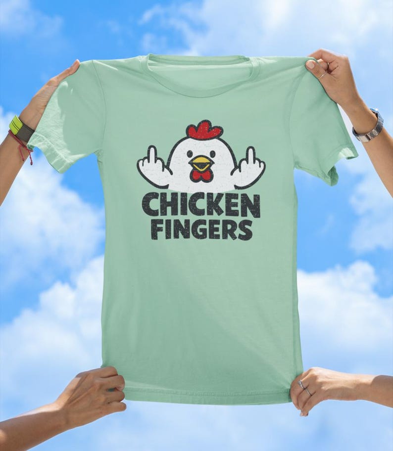 May include: A light green t-shirt featuring a graphic of a white chicken with a red comb and yellow beak, holding up its middle fingers on both sides. Below the chicken, the text CHICKEN FINGERS is printed in a bold, distressed black font.
