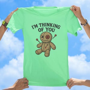May include: A light green t-shirt featuring a voodoo doll graphic and the text "I'm thinking of you".