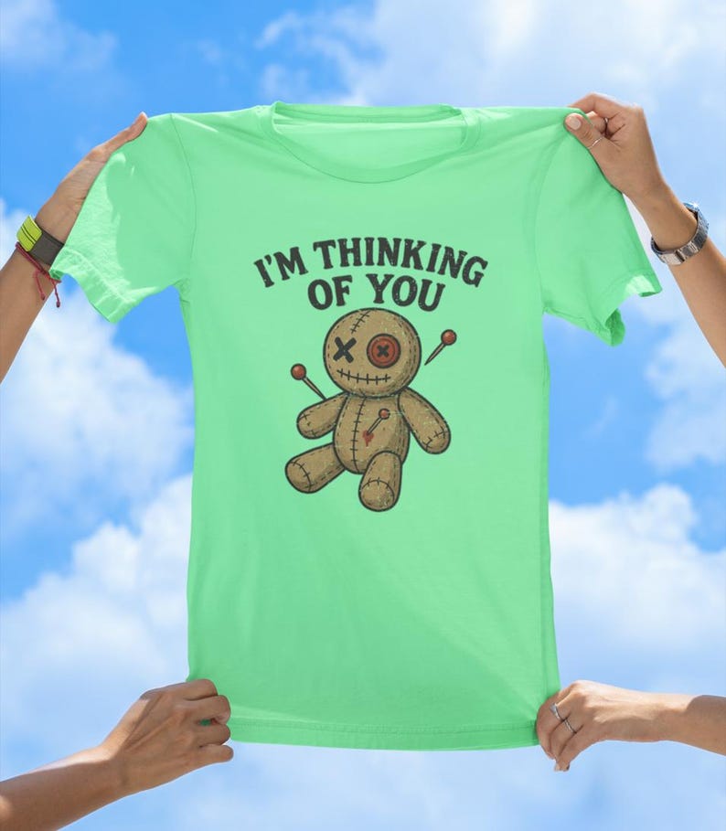 May include: A light green t-shirt featuring a voodoo doll graphic and the text "I'm thinking of you".