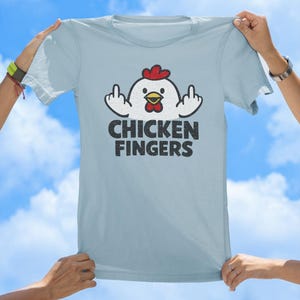 May include: A light blue t-shirt featuring a graphic of a white chicken with a red comb and yellow beak. The chicken is shown with its wings raised, displaying middle fingers. Below the graphic, the text CHICKEN FINGERS is printed in black.