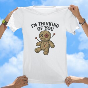 May include: White t-shirt featuring the text "I'm thinking of you" and a cartoon voodoo doll with needles sticking out of it.