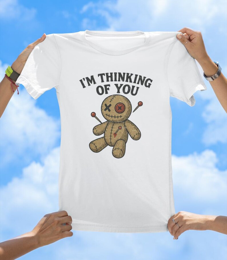 May include: White t-shirt featuring the text "I'm thinking of you" and a cartoon voodoo doll with needles sticking out of it.