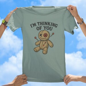 May include: A light blue t-shirt featuring a voodoo doll graphic and the text "I'm thinking of you".