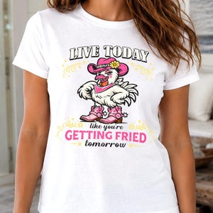 Puede incluir: Una camiseta blanca de manga corta con el gr&aacute;fico de un pollo que lleva un sombrero de vaquero rosa, un pa&ntilde;uelo rosa y botas de vaquero rosas. El texto dice: LIVE TODAY like you're GETTING FRIED tomorrow, con adornos decorativos amarillos.